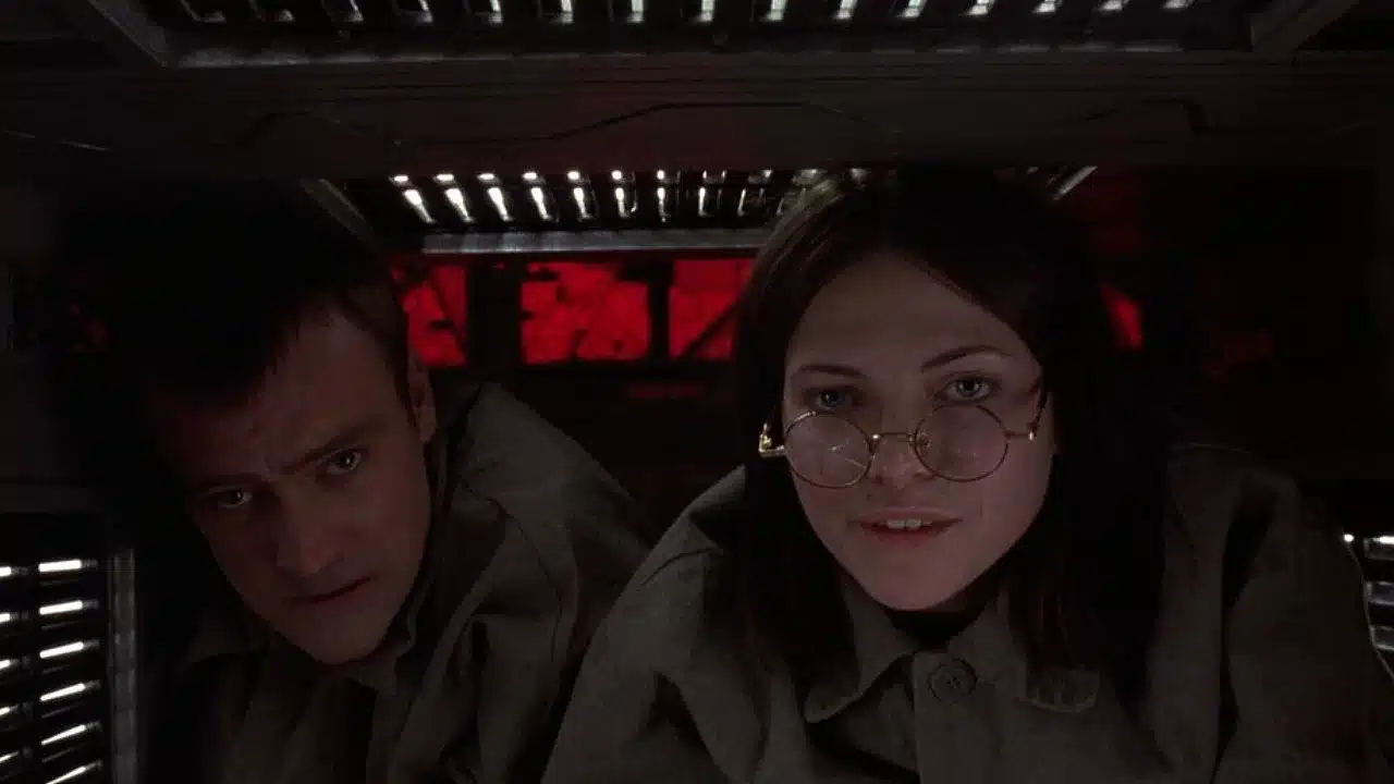 Two stars of CUBE (1997)--one man and one woman in glasses--look directly into the camera in a dark, enclosed space.