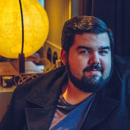 Photo of Allen Longstreet, author of Underbelly (2026). Allen sits in in a dimly lit room in front of a spherical lamp. He has dark, neatly styled hair and a dark beard.