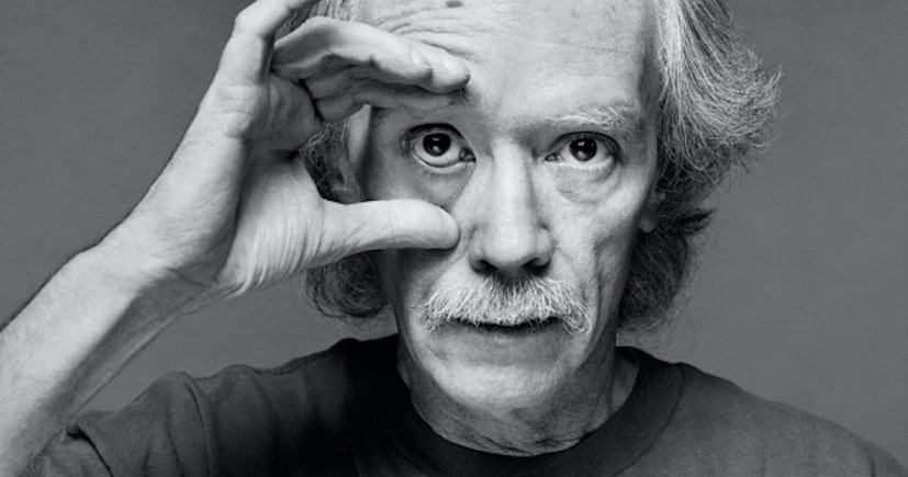 Photo of John Carpenter; black and white photo of older man holding his eye open with his right hand.